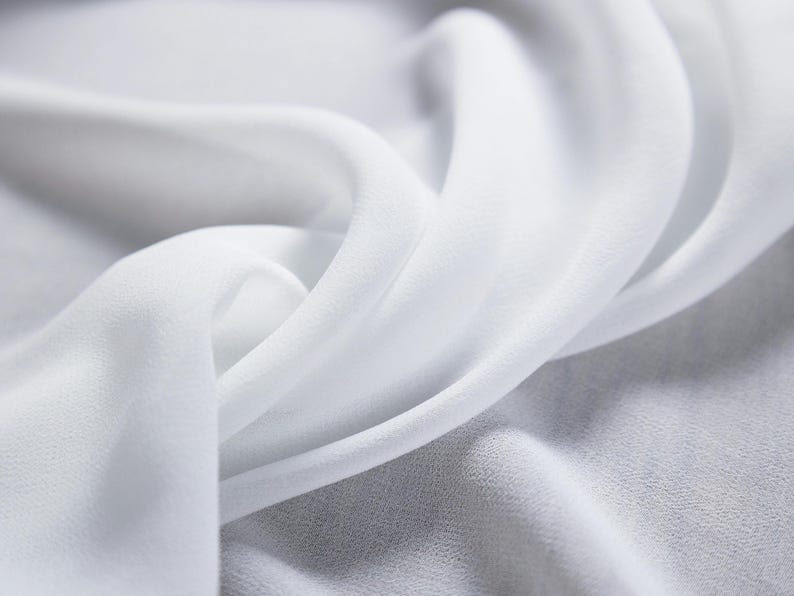 May include: Close-up of flowing white fabric with soft folds and a delicate texture. The fabric appears to be lightweight, possibly silk or chiffon, with a subtle sheen. The image highlights the fabric's smooth surface and gentle curves.