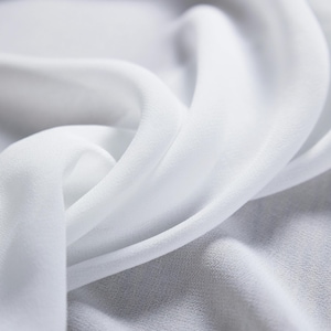 May include: Close-up of flowing white fabric with soft folds and a delicate texture. The fabric appears to be lightweight, possibly silk or chiffon, with a subtle sheen. The image highlights the fabric's smooth surface and gentle curves.