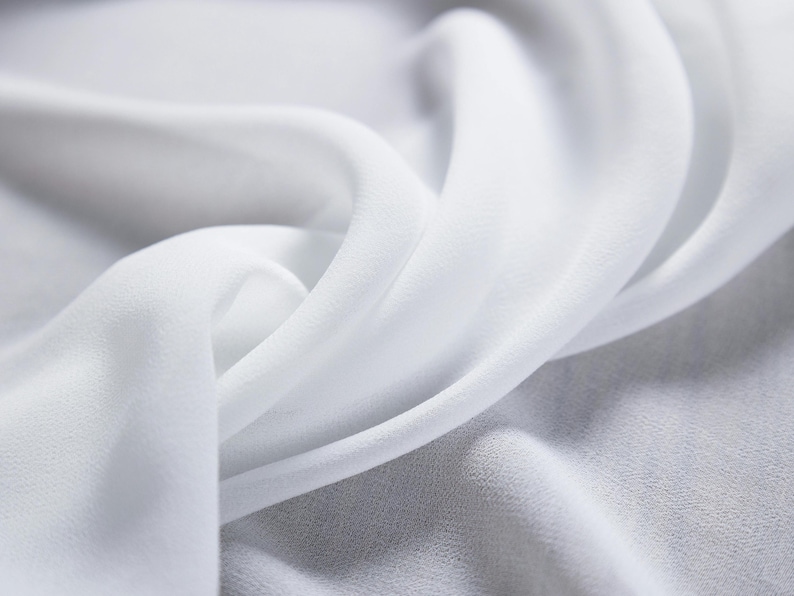 May include: Close-up of flowing white fabric with soft folds and a delicate texture. The fabric appears to be lightweight, possibly silk or chiffon, with a subtle sheen. The image highlights the fabric's smooth surface and gentle curves.