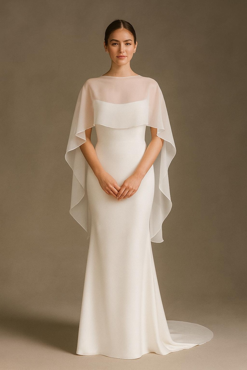 May include: A white wedding dress with a sheer, flowing cape. The dress is a simple, elegant design with a fitted bodice and a long, flowing skirt. The cape adds a touch of ethereal beauty to the bridal gown.