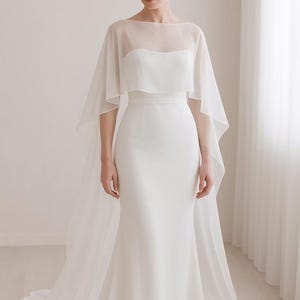 May include: A white wedding dress with a sheer cape overlay. The dress features a fitted bodice and a flowing skirt that flares out at the bottom. The cape drapes over the shoulders and extends to the floor, creating an elegant silhouette.
