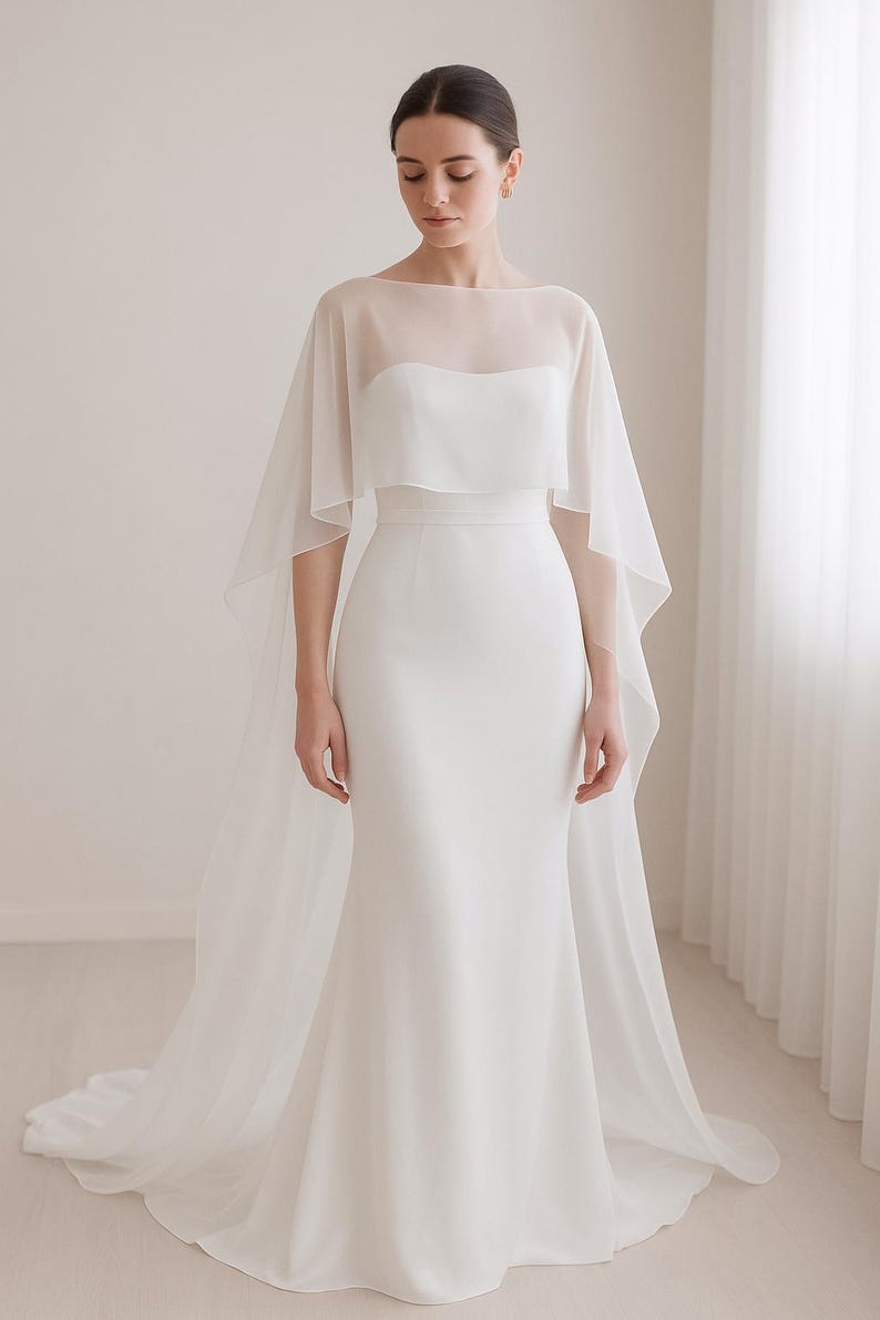 May include: A white wedding dress with a sheer cape overlay. The dress features a fitted bodice and a flowing skirt that flares out at the bottom. The cape drapes over the shoulders and extends to the floor, creating an elegant silhouette.