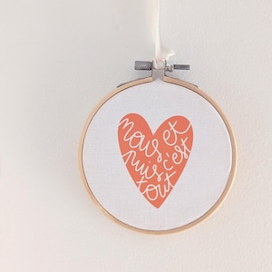 Può includere: Un cerchio da ricamo bianco con un cuore corallo che dice "nous et puis c'est tout" in scrittura corsiva bianca.