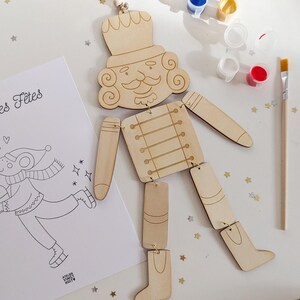 Peut inclure: Un casse-noisette articulé en bois avec une ficelle pour suspendre, ainsi que des pots de peinture et un pinceau. Une page à colorier avec le texte "les Fêtes" est également présente. La figurine est en bois clair.
