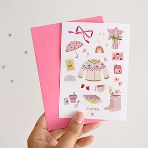 Op de afbeelding: Een roze wenskaart met illustraties met lentethema, waaronder een trui, een paraplu, bloemen en aardbeien. De kaart wordt vastgehouden met een roze envelop. Het woord "mars" staat op de kaart.