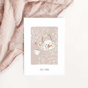 Peut inclure: Carte blanche avec illustration d'une théière et d'une tasse à fleurs. La théière a une anse marron et est ornée de fleurs. Les mots "TEA TIME" sont imprimés en bas. Un tissu rose est en arrière-plan.