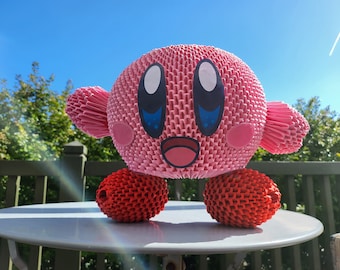 May include: A pink and red origami figure of the video game character Kirby. The figure is made of folded paper and has a round body with large eyes and a small mouth. The figure is sitting on a white table.