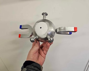 Puede incluir: Un modelo 3D impreso en plata de un Pokémon Magnemite. El modelo tiene dos imanes de herradura plateados unidos a los lados. Los imanes tienen acentos rojos y azules.