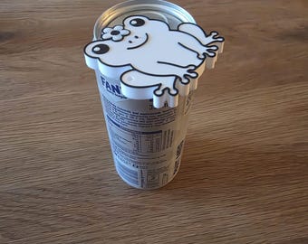 May include: A white plastic drink can topper shaped like a frog with a small flower on its head. The frog has black outlines, black eyes, and a smiling mouth. It is attached to the top rim of a silver aluminum soda can with visible text.