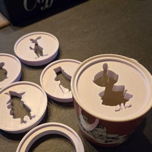 Peut inclure: Une tasse rouge et blanche avec un couvercle orné d'un motif découpé. Plusieurs pochoirs violets clairs avec des formes d'animaux sont éparpillés autour de la tasse. Un coffret en bois noir avec l'inscription "Coffee Bar" est en arrière-plan.