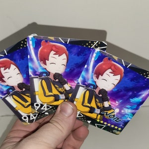Puede incluir: Tres tarjetas coloridas con un personaje de dibujos animados con cabello rojo que lleva una chaqueta amarilla y negra. El personaje sonr&iacute;e y tiene la mano en la barbilla. Las tarjetas tienen un fondo azul y morado con estrellas y un borde blanco. El texto "Nonu" est&aacute; impreso en la parte inferior de una tarjeta.