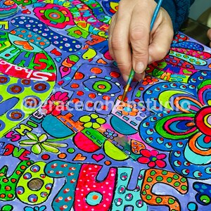 Puede incluir: Una obra de arte vibrante y colorida en progreso, con una mano sosteniendo un pincel. El dise&ntilde;o incluye patrones audaces y caprichosos, flores y texto como "LOVE" y "SHALOM" en tonos brillantes sobre un fondo p&uacute;rpura. El artista est&aacute; agregando detalles.