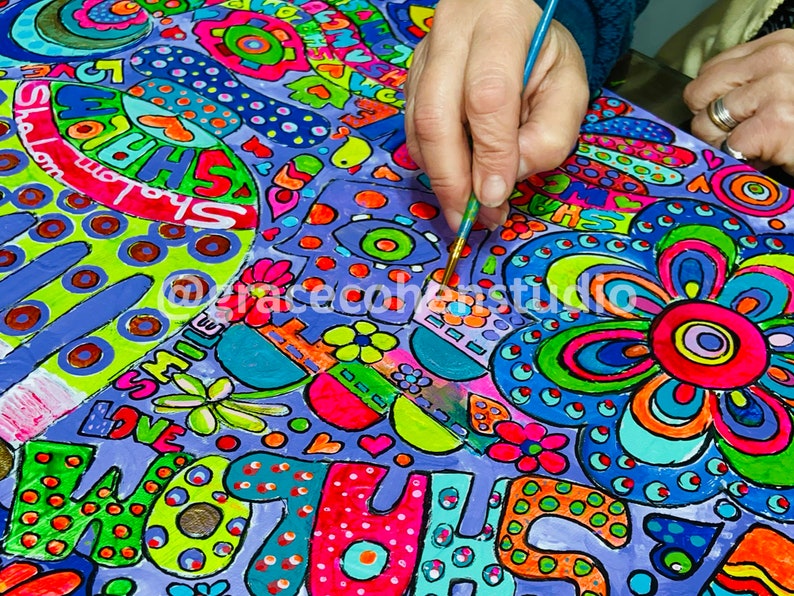 Puede incluir: Una obra de arte vibrante y colorida en progreso, con una mano sosteniendo un pincel. El dise&ntilde;o incluye patrones audaces y caprichosos, flores y texto como "LOVE" y "SHALOM" en tonos brillantes sobre un fondo p&uacute;rpura. El artista est&aacute; agregando detalles.