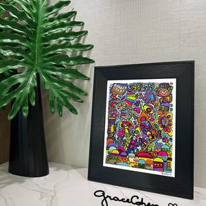 Puede incluir: Una colorida impresi&oacute;n art&iacute;stica enmarcada con la palabra "Shalom" y varias ilustraciones caprichosas. La obra de arte est&aacute; en un marco negro y se muestra sobre una superficie blanca. Una gran hoja verde y un jarr&oacute;n negro est&aacute;n en el fondo.
