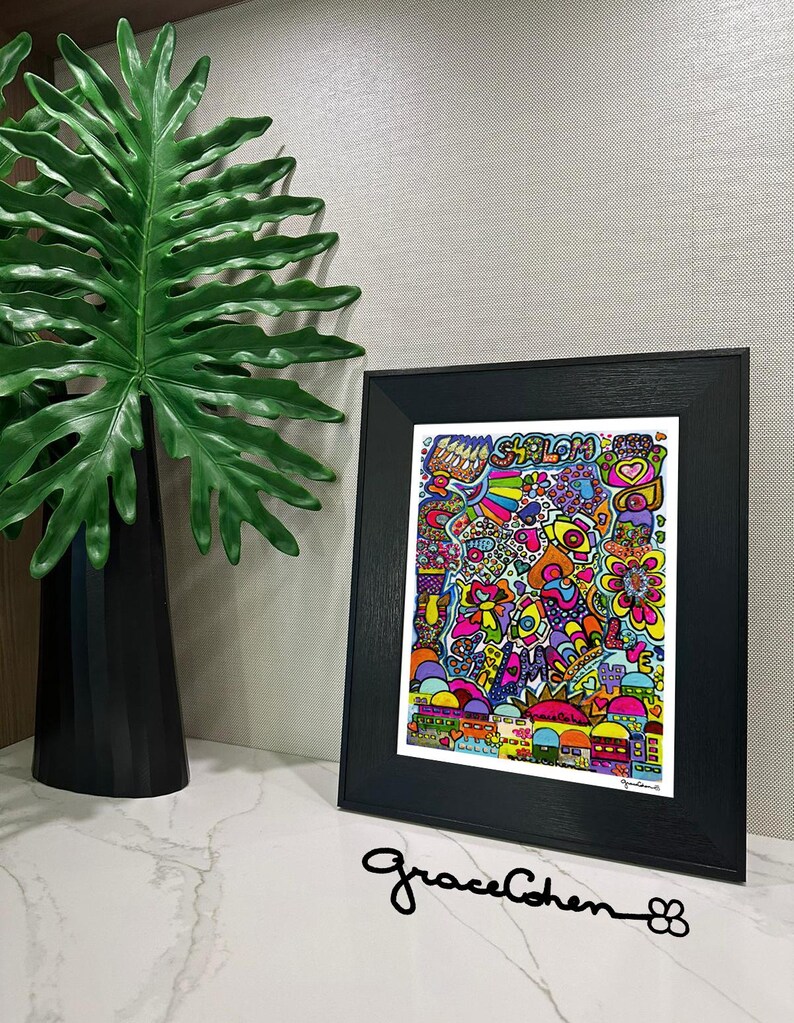 Puede incluir: Una colorida impresi&oacute;n art&iacute;stica enmarcada con la palabra "Shalom" y varias ilustraciones caprichosas. La obra de arte est&aacute; en un marco negro y se muestra sobre una superficie blanca. Una gran hoja verde y un jarr&oacute;n negro est&aacute;n en el fondo.