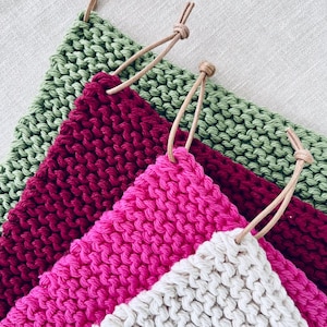 Puede incluir: Cuatro agarraderas de olla de punto con lazos de cuero. Las agarraderas de olla son en tonos de verde, burdeos, rosa y blanco.