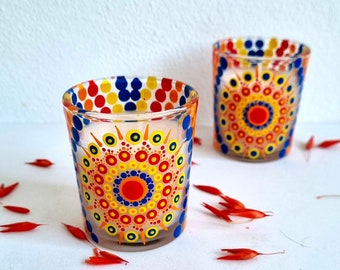 May include: Two glass candle holders with a colorful mandala design. The holders are painted with red, yellow, blue, and white dots. The holders are on a white surface with red flower petals.