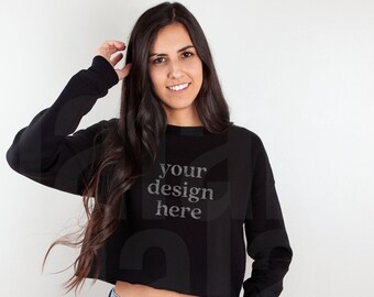 May include: A black cropped sweatshirt with long sleeves and the text "your design here" in a light grey color. The sweatshirt is paired with light blue denim jeans.