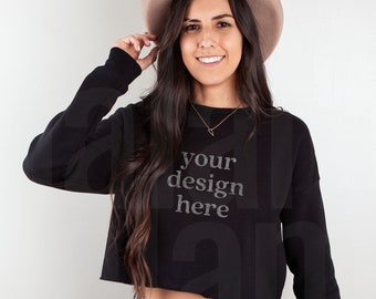 May include: Black cropped sweatshirt with the text "your design here". The sweatshirt is paired with light blue jeans and a tan hat. The model is smiling and wearing a gold necklace.