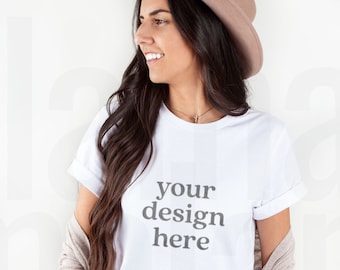 Puede incluir: Una mujer con una camiseta blanca con el texto "your design here" impreso en la parte delantera. También lleva un sombrero marrón y unos vaqueros azules.