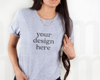 Può includere: Una maglietta grigio chiaro con maniche arrotolate. Il testo "your design here" è stampato in grigio scuro sul davanti. La modella indossa pantaloni neri e gioielli d'oro.