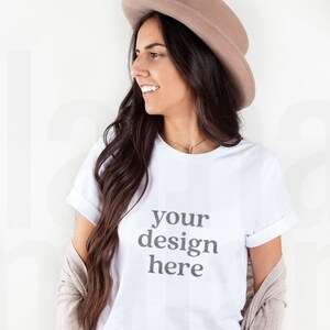 May include: A woman wearing a white t-shirt with the text "your design here" printed on the front. She is also wearing a brown hat and blue jeans.