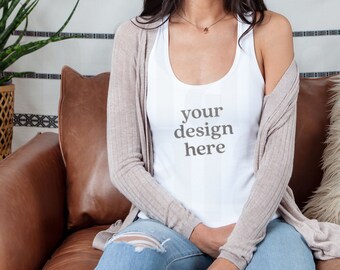 Pode incluir: Top branco com a frase "your design here". O top é usado com um cardigan castanho claro e jeans azuis. A pessoa está sentada num sofá de couro castanho.