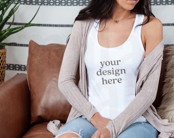 Pode incluir: Uma mulher vestindo um top branco sem mangas com o texto "your design here" nele. Ela está sentada em um sofá de couro marrom e usando um cardigã marrom claro e jeans azul.