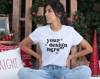 May include: A light gray t-shirt with the text "your design here" in black. The model is wearing blue jeans and sitting in front of a wooden backdrop decorated with Christmas ornaments and a pillow that says "BRIGHT."