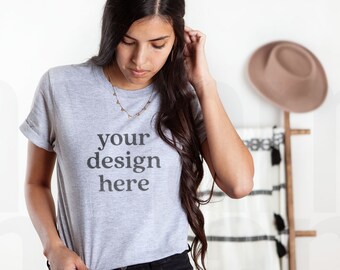 Può includere: Una maglietta grigio chiaro con maniche arrotolate e la scritta "your design here" in un carattere grigio scuro. La maglietta è abbinata a jeans neri. È visibile una collana d'oro. Un cappello e una scala decorativa sono sullo sfondo.
