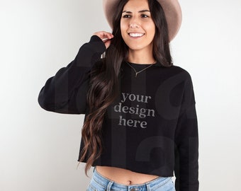 May include: A black cropped sweatshirt with long sleeves and the text "your design here" printed on the front. The model is wearing blue jeans and a brown hat.