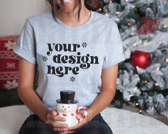 May include: A light gray t-shirt with the text "your design here" in black, with snowflake accents. The person is holding a snowman-shaped mug with a black top hat and a red and white striped scarf. A Christmas tree is in the background.