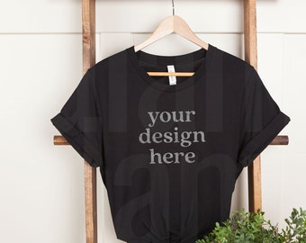 Puede incluir: Una camiseta negra con las mangas remangadas colgada de una escalera de madera. La camiseta tiene el texto "your design here" impreso en la parte delantera.