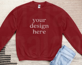 May include: A maroon sweatshirt with the text "your design here" in a distressed font. Light blue jeans are folded at the bottom right. The sweatshirt is on a neutral-toned woven surface.
