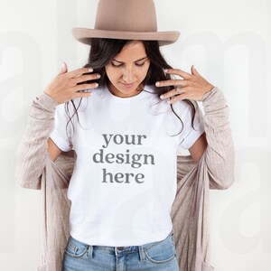 Puede incluir: Una camiseta blanca con el texto "your design here" se usa con vaqueros azules y una rebeca beige. Un sombrero beige est&aacute; sobre la cabeza de la persona. La imagen es una maqueta de producto, ideal para mostrar dise&ntilde;os de camisetas personalizados.