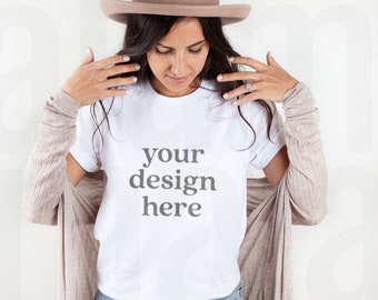 Puede incluir: Una camiseta blanca con el texto "your design here" se usa con vaqueros azules y una rebeca beige. Un sombrero beige está sobre la cabeza de la persona. La imagen es una maqueta de producto, ideal para mostrar diseños de camisetas personalizados.