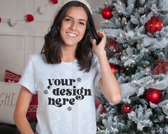 May include: A light gray t-shirt with the text "your design here" in a black, decorative font. The person is wearing a white knit cap and black leggings. A decorated Christmas tree with red ornaments is in the background.