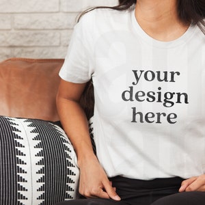 May include: A white t-shirt with the text "your design here" printed on the front. The t-shirt is being worn by a person sitting on a brown leather couch.