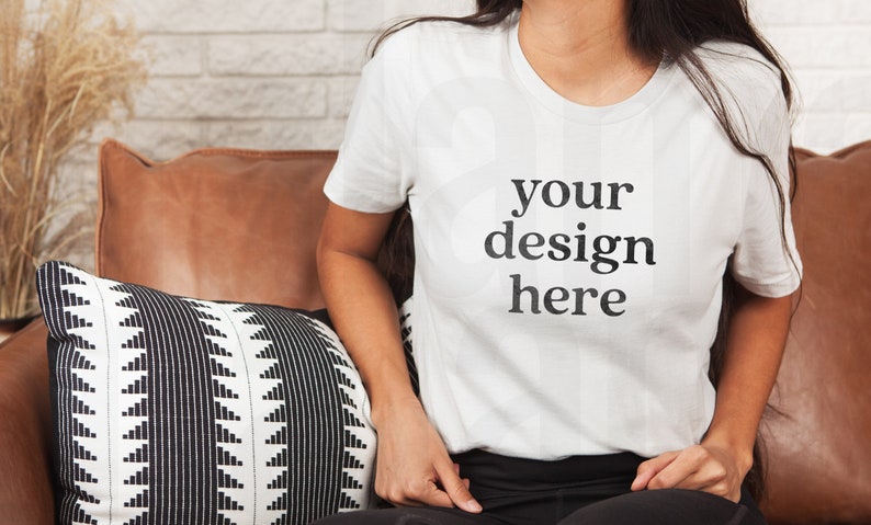 May include: A white t-shirt with the text "your design here" printed on the front. The t-shirt is being worn by a person sitting on a brown leather couch.