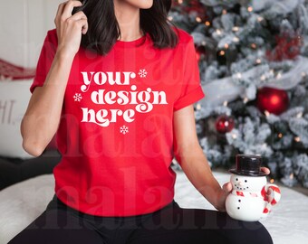 Könnte beinhalten: Ein rotes T-Shirt mit dem weißen Text "your design here", verziert mit Schneeflocken. Die Person hält einen schneemannförmigen Becher mit schwarzem Zylinder und einem Zuckerstangen-Griff. Im Hintergrund ist ein Weihnachtsbaum zu sehen.