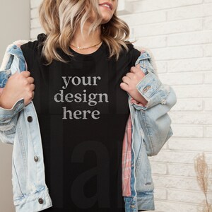 May include: A black t-shirt with the text "your design here" in a distressed white font. The t-shirt is being worn by a person with blonde hair and a denim jacket.