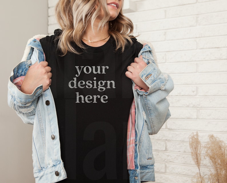 May include: A black t-shirt with the text "your design here" in a distressed white font. The t-shirt is being worn by a person with blonde hair and a denim jacket.