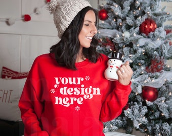 May include: A woman wearing a red sweatshirt with the text "your design here" in white, sitting in front of a decorated Christmas tree. She is holding a white snowman mug.
