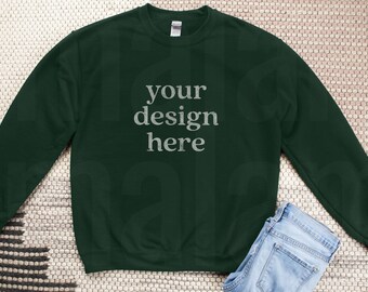 May include: A dark green sweatshirt with the text "your design here" in a light grey color. A pair of light blue jeans is partially visible in the bottom right corner. The sweatshirt is displayed on a neutral-toned woven surface.