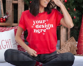 May include: A red t-shirt with the text "your design here" in white, with snowflake accents. The person is sitting cross-legged in front of a Christmas-themed backdrop, with a pillow that says "BRIGHT."
