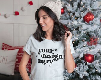 May include: A light gray t-shirt with the text "your design here" in a black, decorative font. The model is wearing a cream-colored knit hat. A Christmas tree with red ornaments and white lights is in the background.