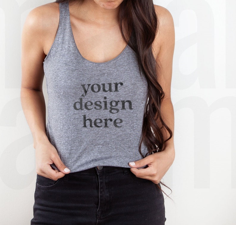 May include: A heather gray tank top with the text "your design here" printed on the front. The tank top has a scoop neck and is paired with black jeans. The model is holding the bottom of the tank top.