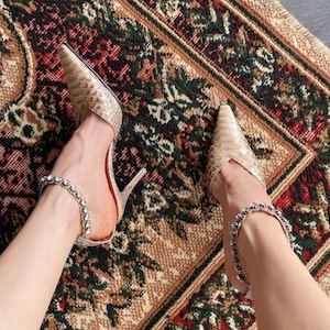 May include: Tan snakeskin-effect high heels with pointed toes and a delicate ankle strap embellished with small, sparkling rhinestones. The shoes are set against a patterned rug with a red, green, and beige colour scheme.