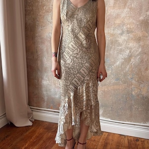 May include: A sleeveless, floor-length dress in a champagne color with a floral pattern. The dress features a V-neckline and a ruffled hem. The model is wearing burgundy heels.