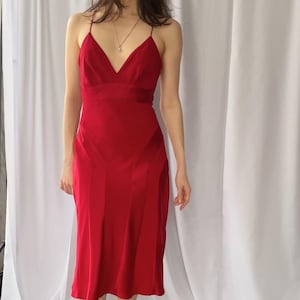 Può includere: Un vivace abito midi rosso con spalline sottili e scollo a V. L'abito ha un corpetto aderente e una gonna fluida. La modella indossa tacchi neri. L'abito arriva approssimativamente al ginocchio.